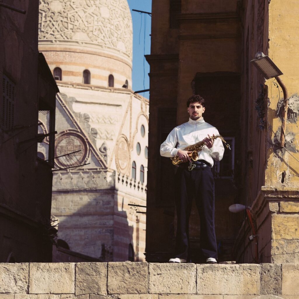 A man in a white shirt and dark pants stands on a stone ledge holding a stringed instrument, evoking a saint Levant vibe, with ornate historic buildings and a large domed structure in the background under daylight.