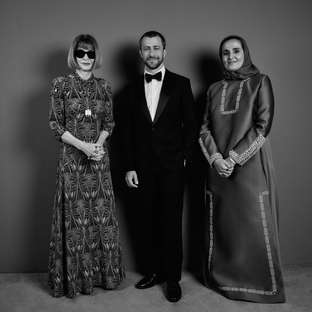 Three people stand side by side against a plain backdrop at the Franca Fund Gala: a woman in a patterned dress and sunglasses, a man in a tuxedo, and a woman in an ornate long dress and headscarf. The photo is black and white.