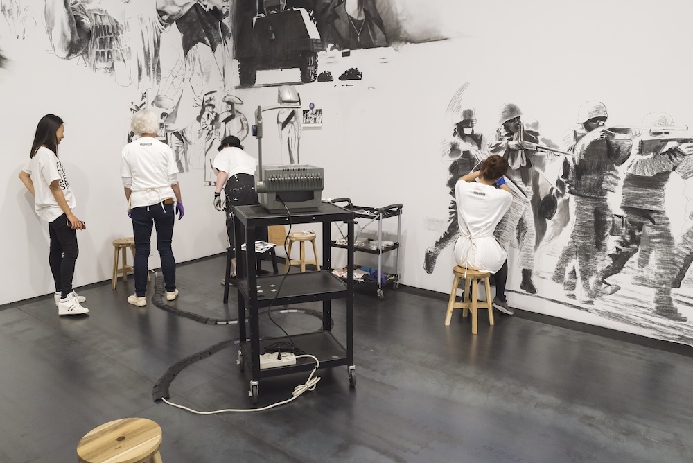 Four people work on a detailed black-and-white mural depicting historical scenes, using projectors and stools in a gallery-like space reminiscent of Rirkrit Tiravanija's participatory art. Art supplies and equipment rest on a black cart in the center.