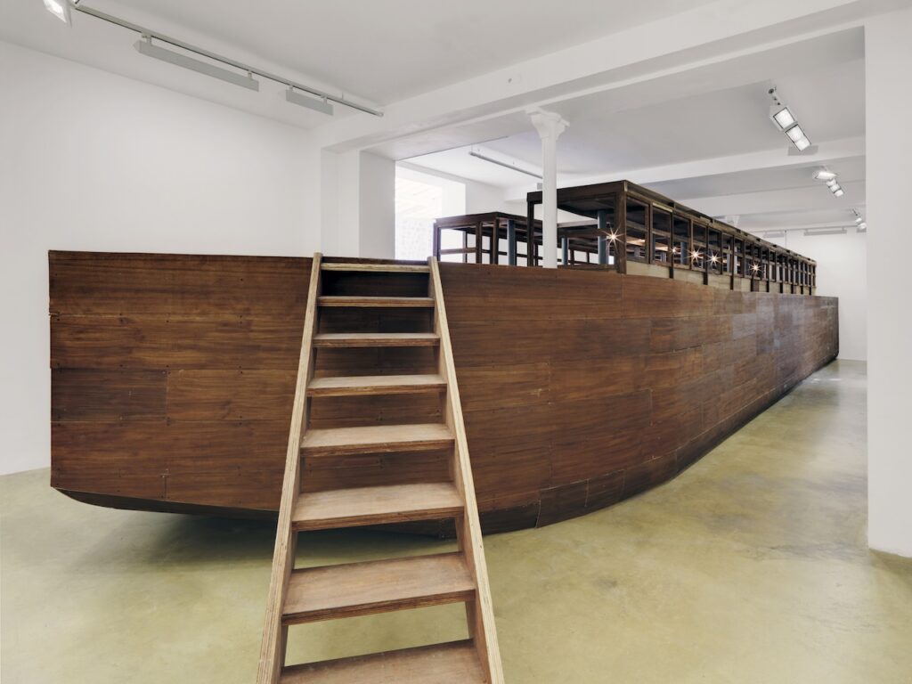 Large wooden structure resembling a boat fills a white-walled room; reminiscent of Rirkrit Tiravanija’s participatory installations, a wooden staircase leads up to its deck, which has benches and railings under bright ceiling lights.