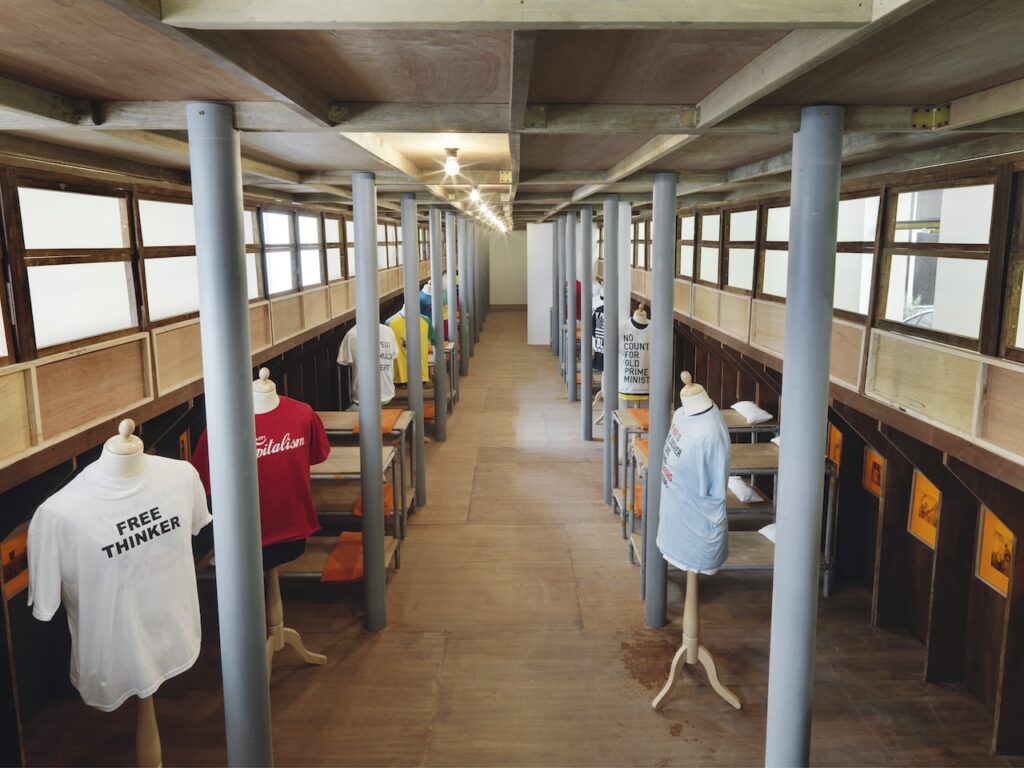 Interior of a wooden building with columns, mannequins wearing various printed T-shirts—reminiscent of Rirkrit Tiravanija's communal art spaces—displayed on either side, and natural light streaming through large side windows.