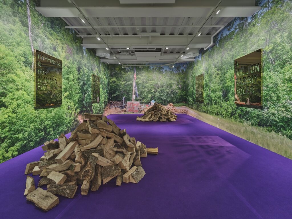 An art gallery room inspired by Rirkrit Tiravanija features walls covered in forest imagery, mirrored text panels, and piles of chopped wood on a purple carpet. The white ceiling is lit brightly, with a forest scene visible at the far end.
