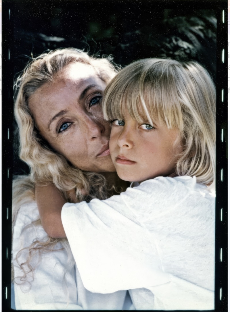 A woman with long, wavy blonde hair embraces a young child with straight blonde hair. Both are wearing white shirts and looking at the camera with calm, serious expressions during the Franca Fund Gala.