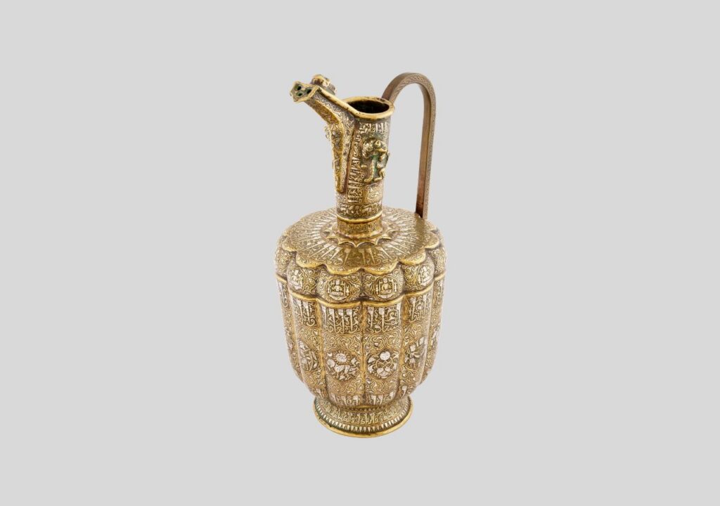 Ornate golden pitcher with intricate floral and geometric patterns evokes the elegance of an empire of light, featuring a curved handle and a detailed spout topped with a small decorative figure. The background is plain and light gray.