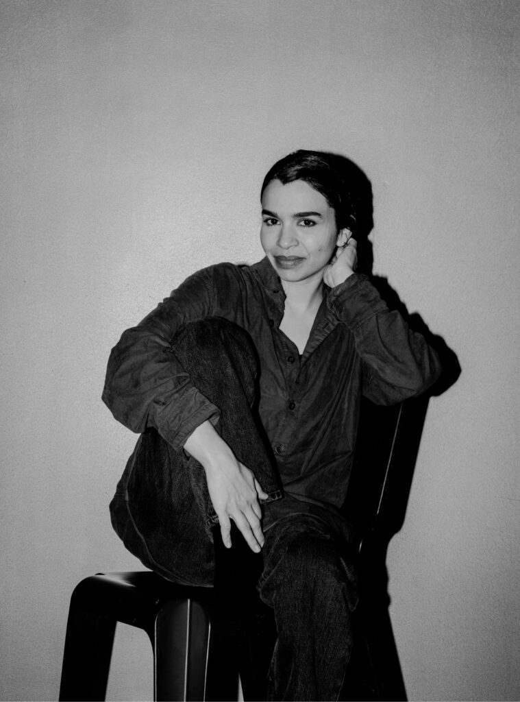 A person with dark hair, wearing a button-up shirt and jeans, sits on a chair with one knee up, resting their arm on it. Reminiscent of Rirkrit Tiravanija’s candid style, they look at the camera and smile slightly in this black-and-white photo.