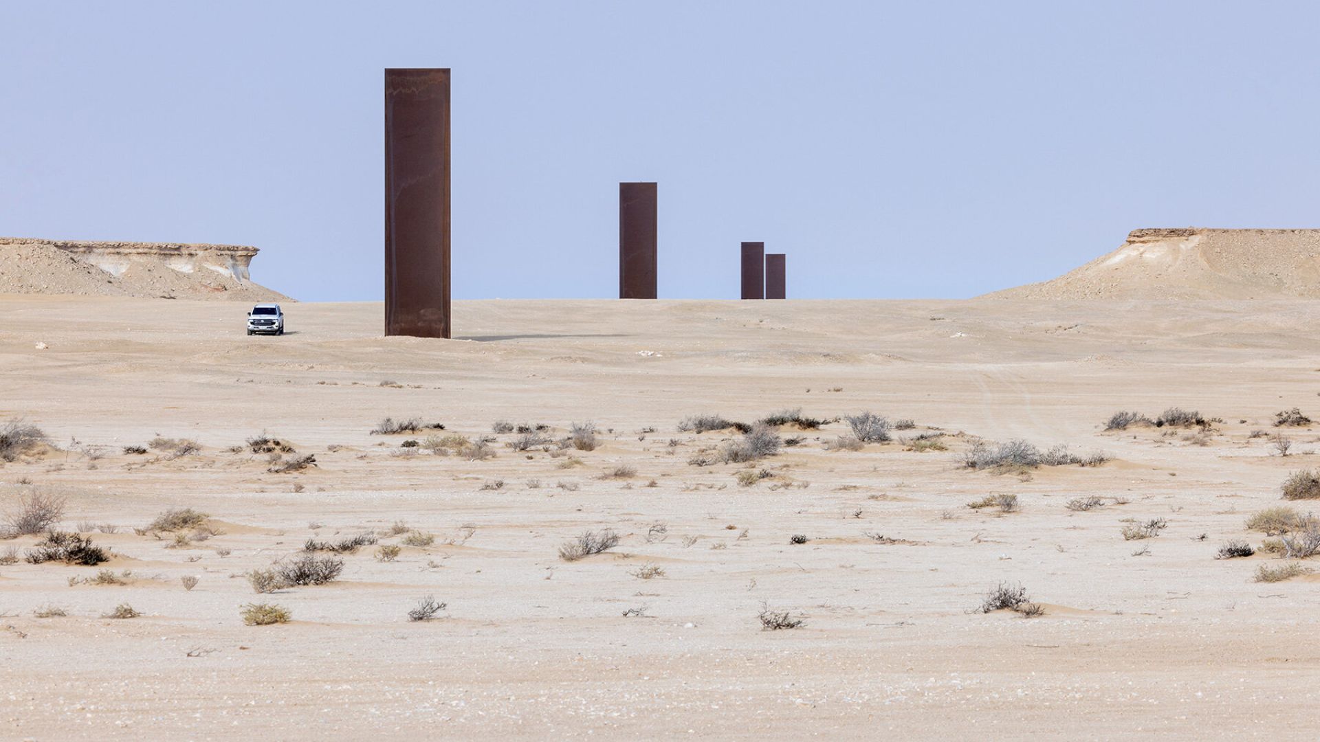 A desert landscape with sparse vegetation, featuring a row of tall, rust-colored rectangular metal pillars—striking examples of public art—and a white vehicle parked near the first pillar under a clear sky.