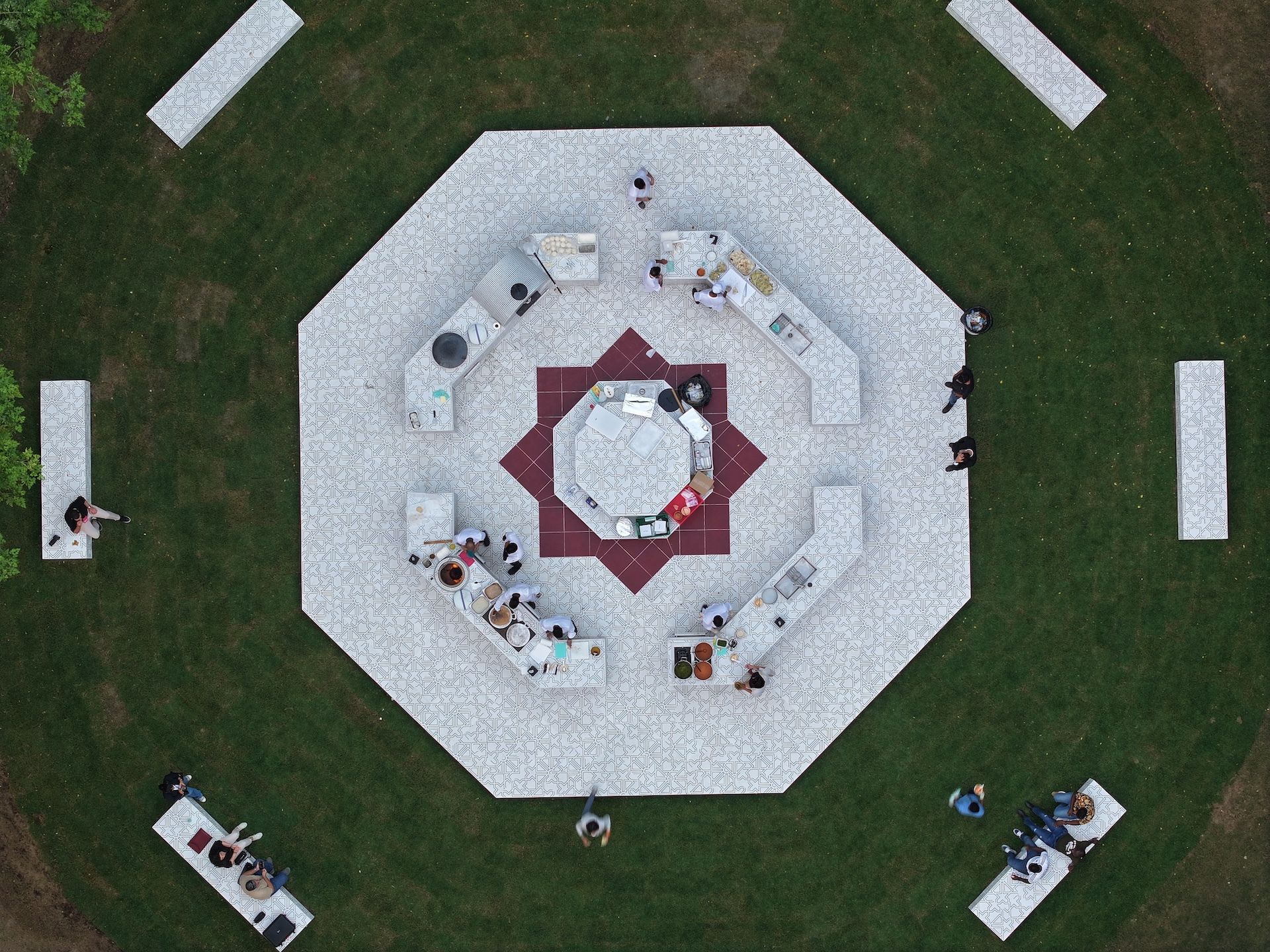 Aerial view of an octagonal outdoor dining area, reminiscent of Rirkrit Tiravanija’s communal installations, with people seated around tables on a patterned surface, surrounded by green grass and white benches.