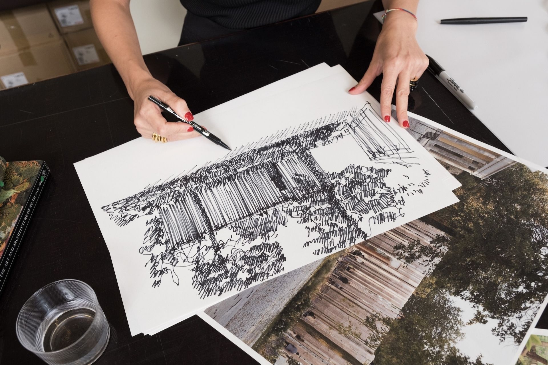 A person with red nail polish sketches a detailed architectural drawing of a building and trees in black ink, referencing printed photos of a wooden structure on a dark table. A glass of water and books are nearby.
