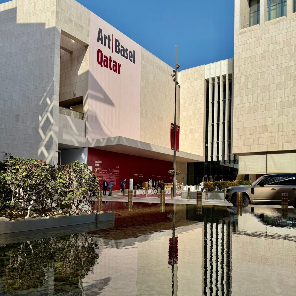 A modern building with Art Basel Qatar signage, people gathering near the entrance, red wall, and a reflecting water feature. A car is parked on the right under a clear blue sky, capturing the vibrant atmosphere of Art Basel Qatar.