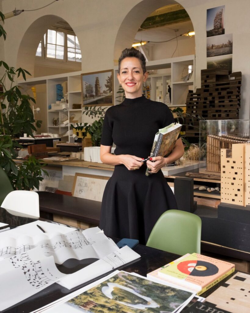 A woman with short hair wearing a black dress stands smiling in a bright, modern studio filled with architectural models, books, and design materials—evoking the creative spirit of Lina Ghotmeh.