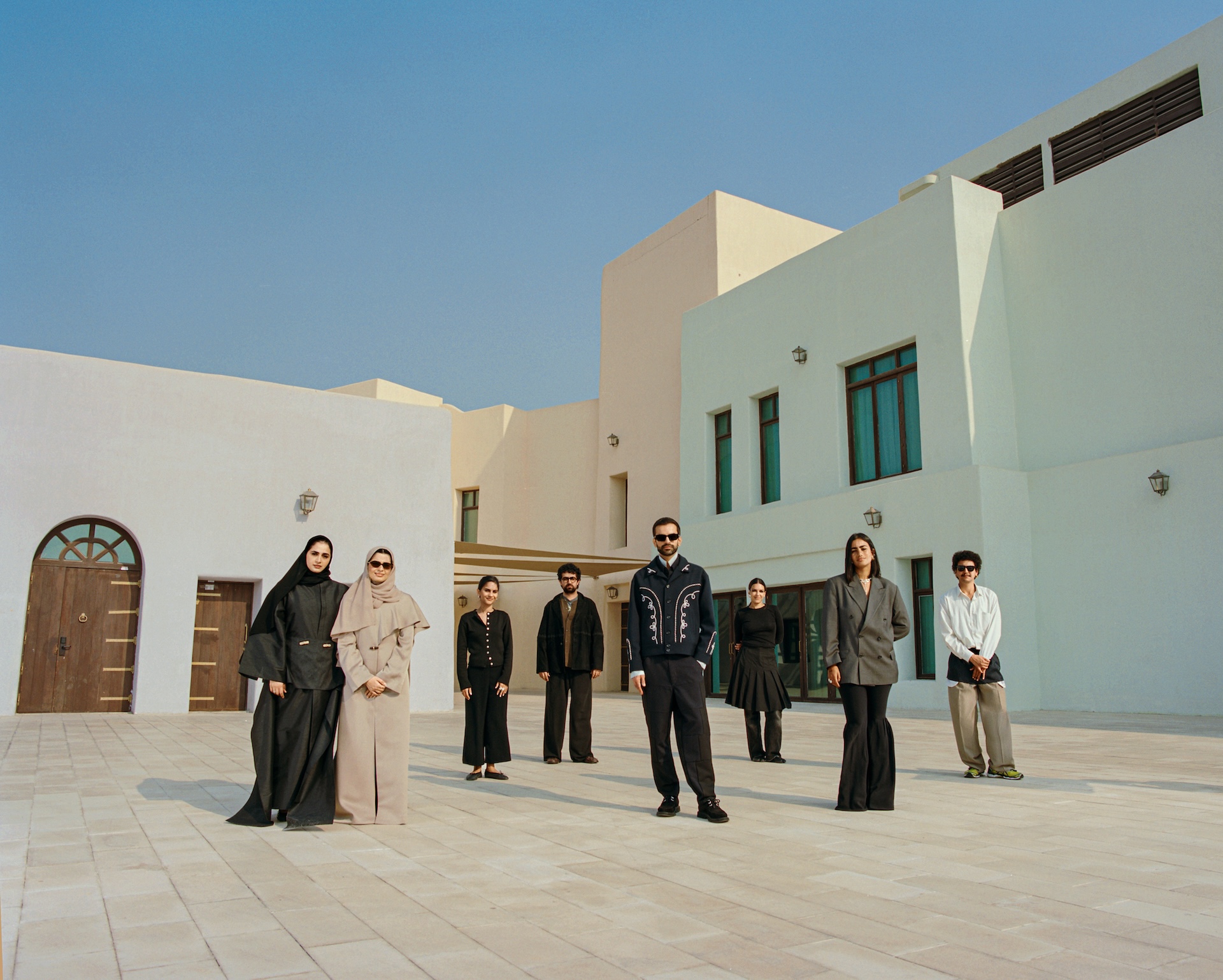 A group of eight people stands spaced apart on a sunlit courtyard with modern white buildings in the background, all wearing modest, stylish clothing and sunglasses, embodying the elegance seen at the Fashion Trust Arabia Prize, under a clear blue sky.