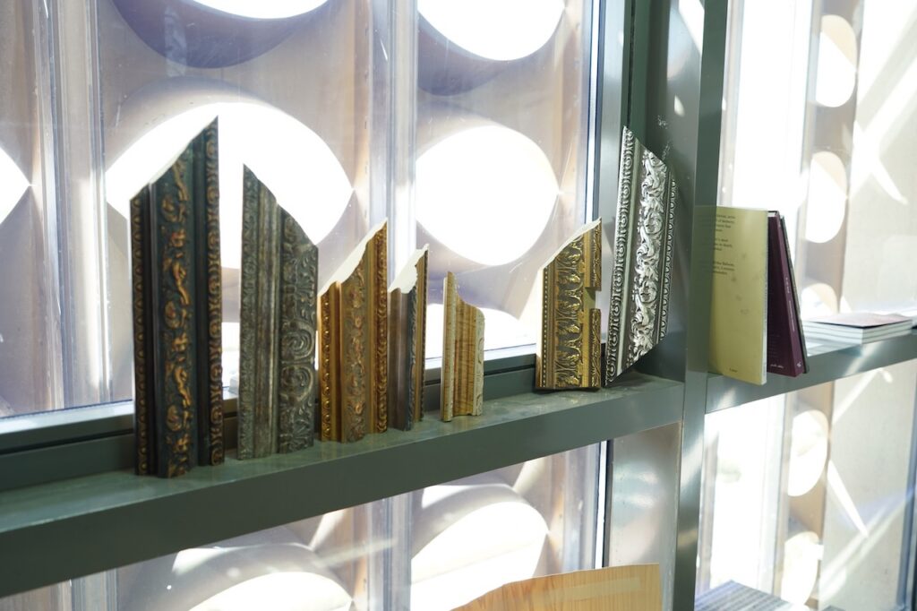 Several ornate, decorative picture frame corners are displayed upright on a window ledge alongside a few stacked books, with sunlight streaming through patterned glass in the background—an arrangement reminiscent of Alla Abdunabi’s artistic style.