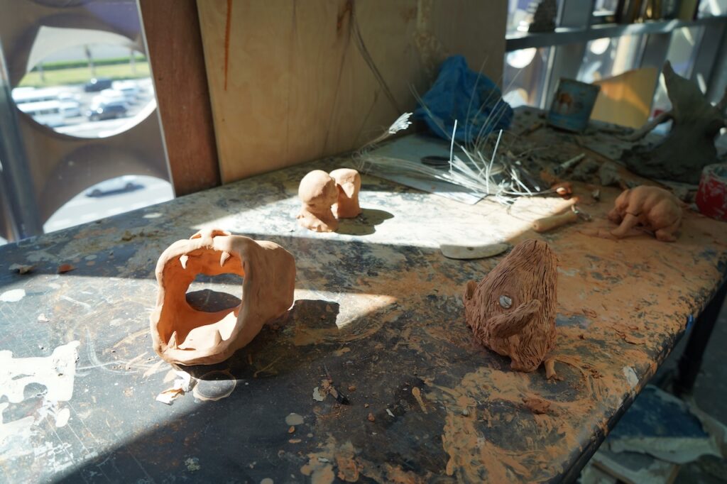 A sunlit worktable scattered with clay dust and tools displays several small, unfinished clay sculptures by Alla Abdunabi and a hollow, rounded clay piece in progress. Sunlight streams in from a nearby window.