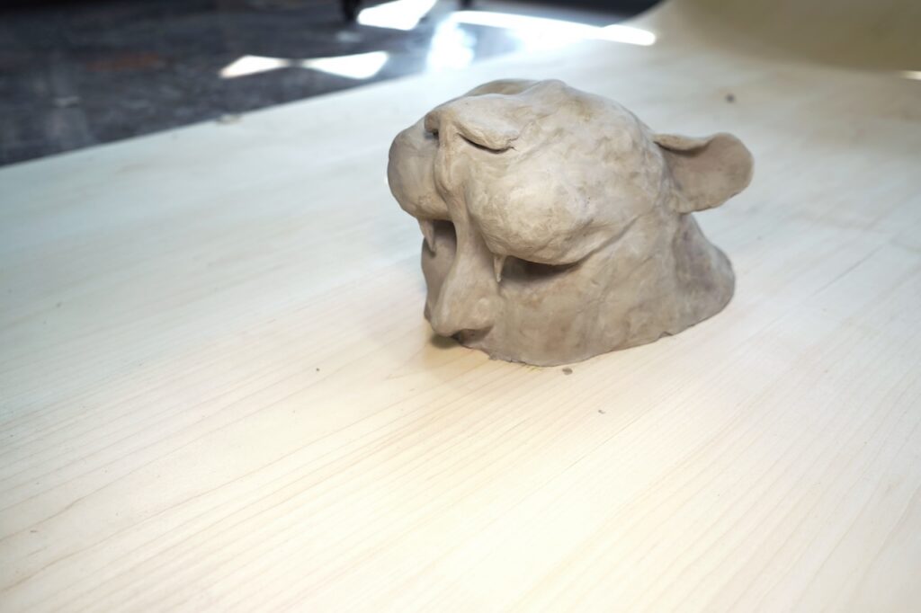 A small clay sculpture by Alla Abdunabi, possibly of a big cat’s head, sits on a light wooden surface. The figure features prominent cheeks, closed eyes, and rounded ears.