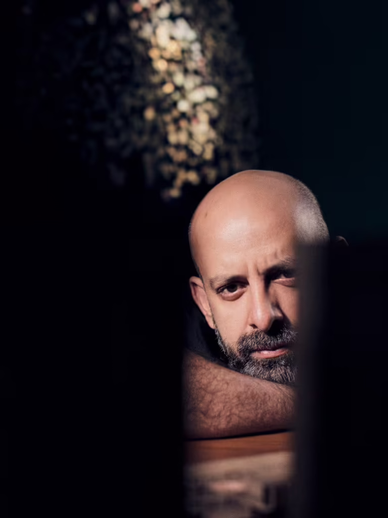A bald man with a beard rests his arms on a surface and looks thoughtfully ahead, as if reflecting on the masterpieces of the Lawh Wa Qalam Museum, with soft lighting highlighting his face against a dark background.