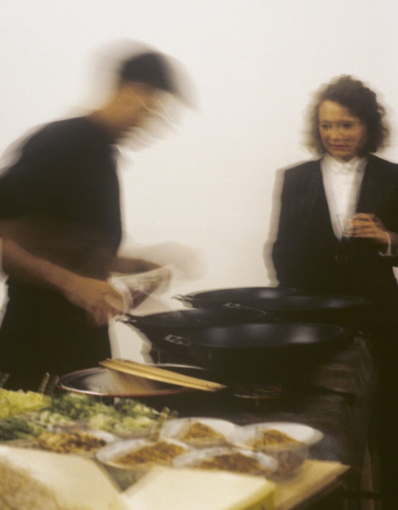 A blurred person is cooking at a table with woks and various ingredients, reminiscent of a Rirkrit Tiravanija installation, while a woman in a suit stands nearby holding a drink and looking on with a concerned expression.