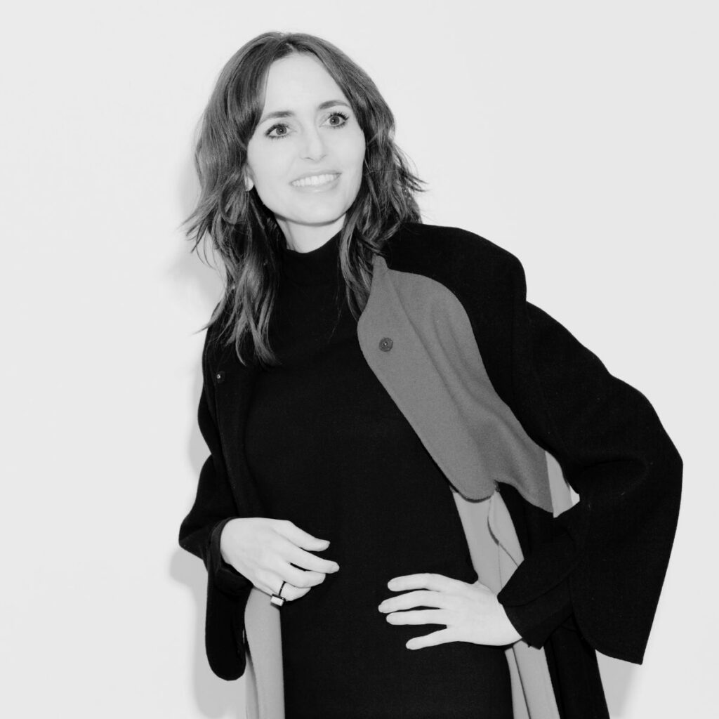 Shaikha Al Sulaiti, with wavy, shoulder-length hair, smiles while wearing a long, dark coat over a black turtleneck dress. She stands against a plain, light background with one hand on her hip.