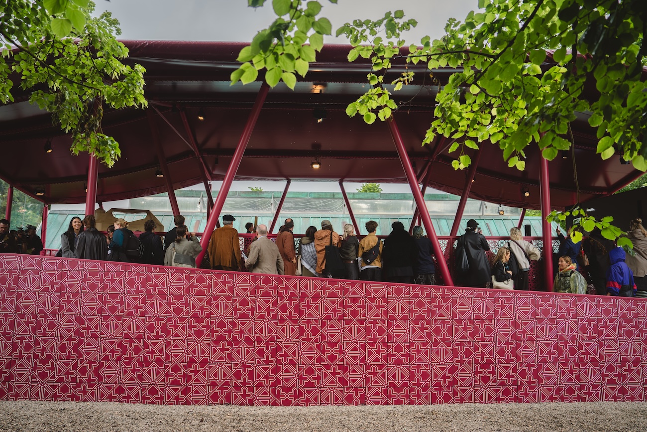 A crowd gathers under a maroon, open-air pavilion with geometric patterns on the wall. Green trees frame the scene, and gravel covers the ground—an elegant setting reminiscent of a Fashion Trust Arabia Prize event.
