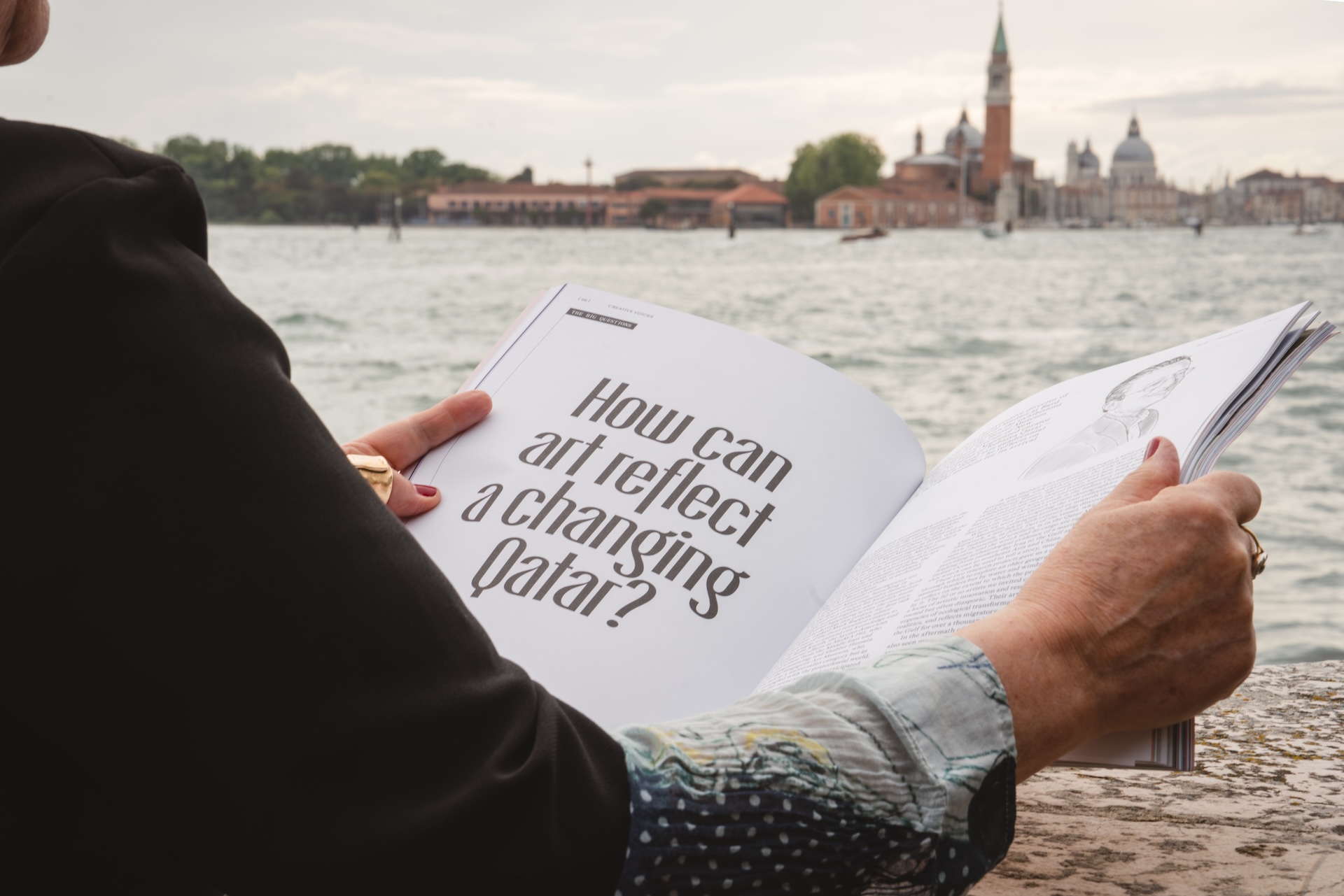 A person holds open a magazine by the water, displaying a page with the headline How can art reflect a changing Qatar? In the background, buildings and a bell tower rise across the water—echoing the creativity of the Fashion Trust Arabia Prize.