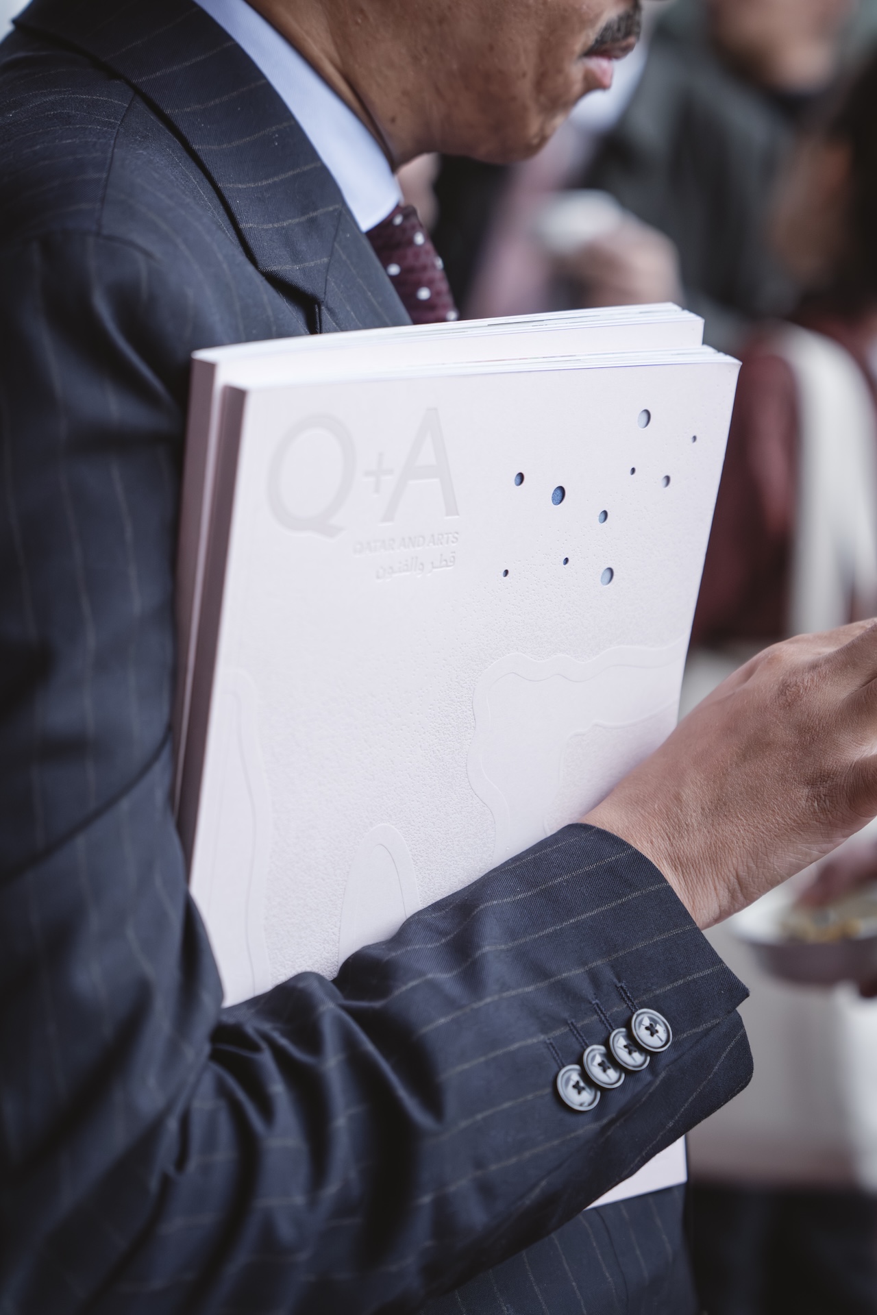 A man in a pinstripe suit holds a large white magazine labeled Q+A under his arm, perhaps awaiting the Fashion Trust Arabia Prize event. Only his midsection and hand are visible, with a blurred background showing people in conversation.