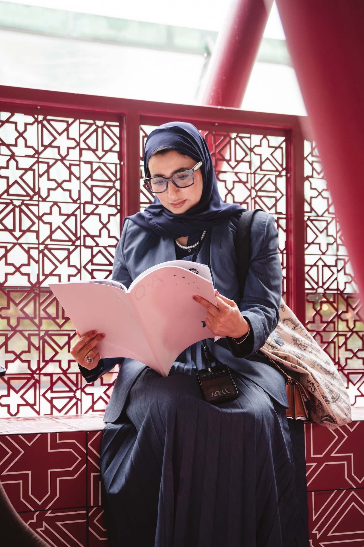 A woman wearing a navy hijab, glasses, and a dark outfit sits on a patterned red bench, reading a document. Sporting a stylish bag and her Fashion Trust Arabia Prize lanyard, she appears focused and engaged in her reading.
