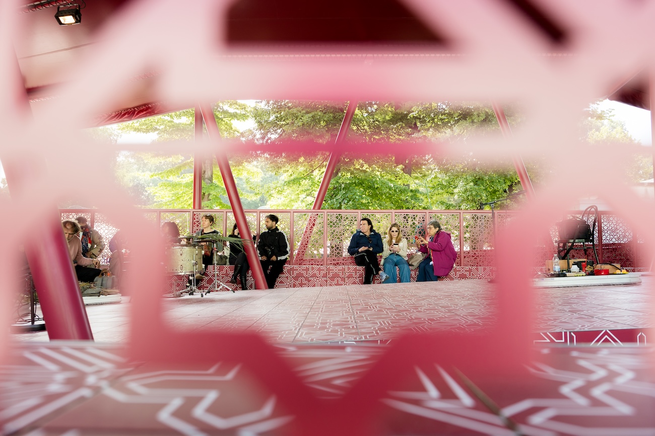 A group of people sits and talks in a colorful outdoor pavilion with geometric pink designs, partially viewed through a decorative screen—a vibrant scene reminiscent of the Fashion Trust Arabia Prize gatherings. Trees and sunlight are visible in the background.