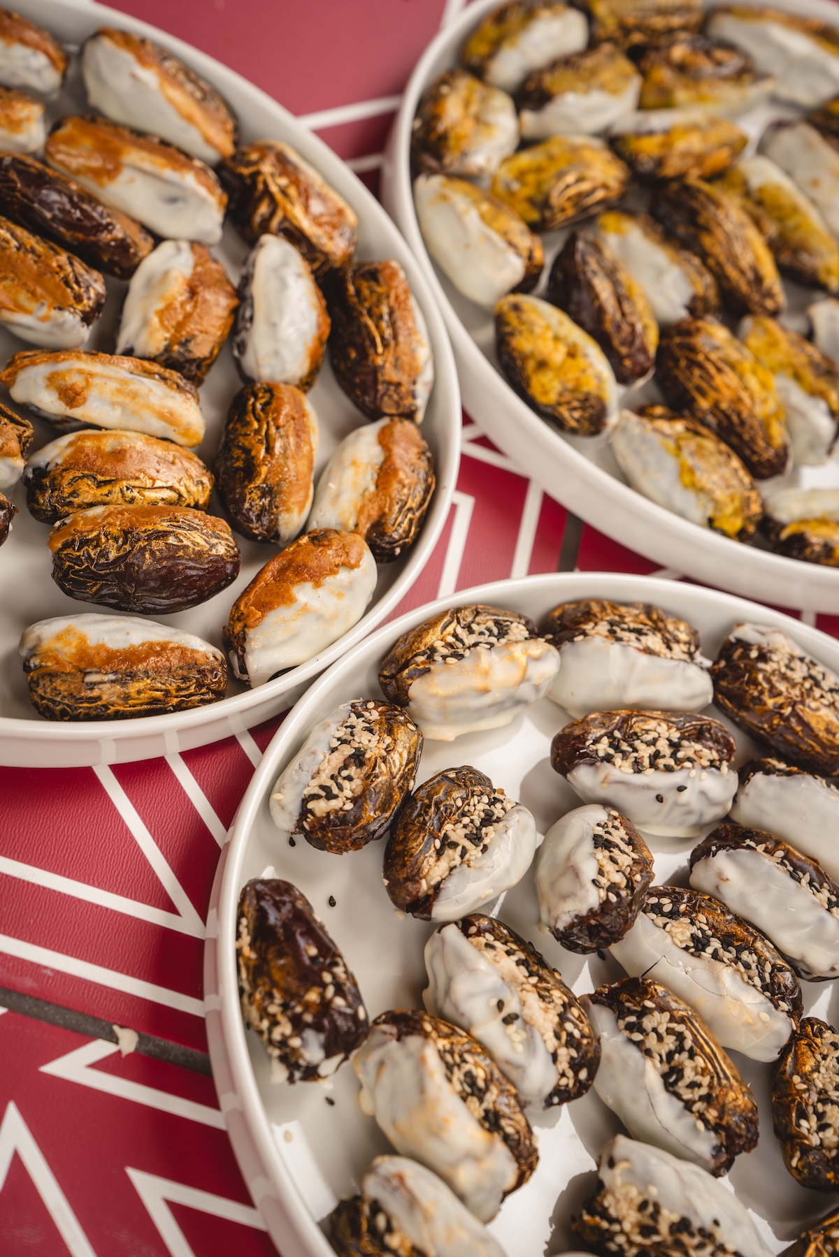 Three plates filled with dates, some partially coated in white icing and sprinkled with nuts or seeds, are arranged on a table with a red and white geometric pattern at the Fashion Trust Arabia Prize event.
