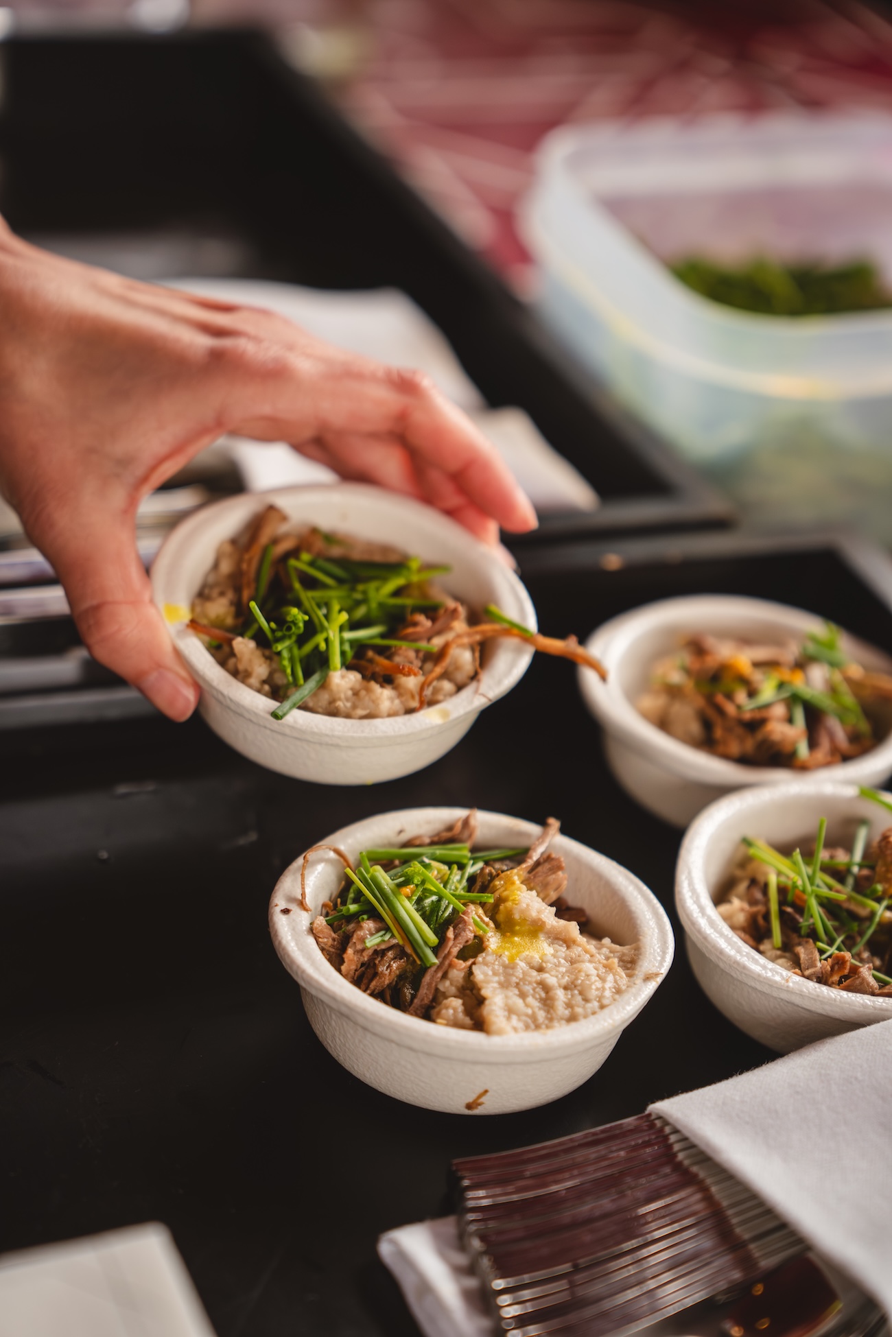 A hand is placing a small white bowl of food garnished with chives next to three similar bowls on a black tray, as if thoughtfully presenting each dish for a Fashion Trust Arabia Prize banquet. The savory meal combines meat and herbs.