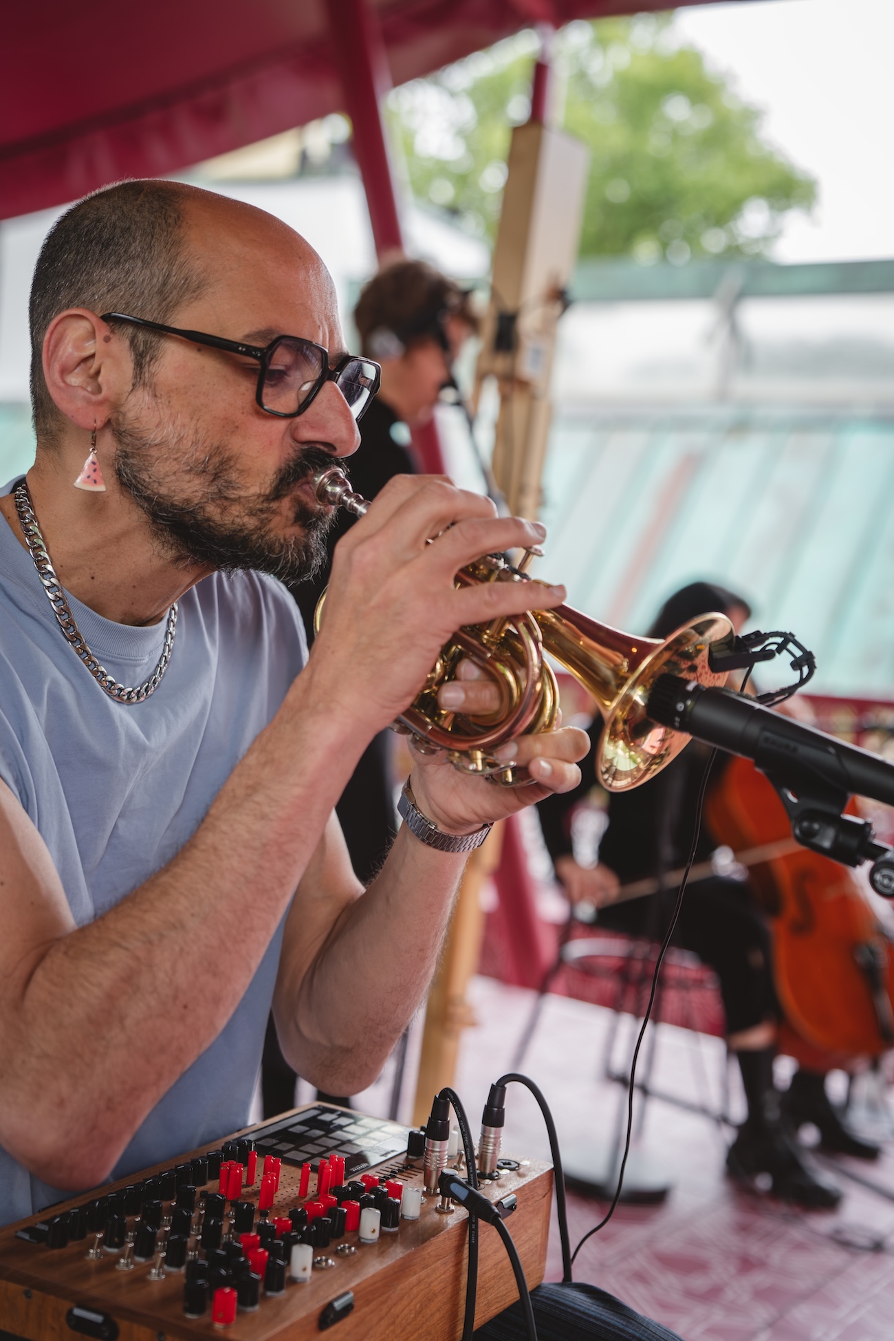 A man wearing glasses and a chain necklace, reminiscent of Fashion Trust Arabia Prize style, plays a small brass horn into a microphone, with a music mixer in front. In the background, two musicians, one with a cello, are also performing.