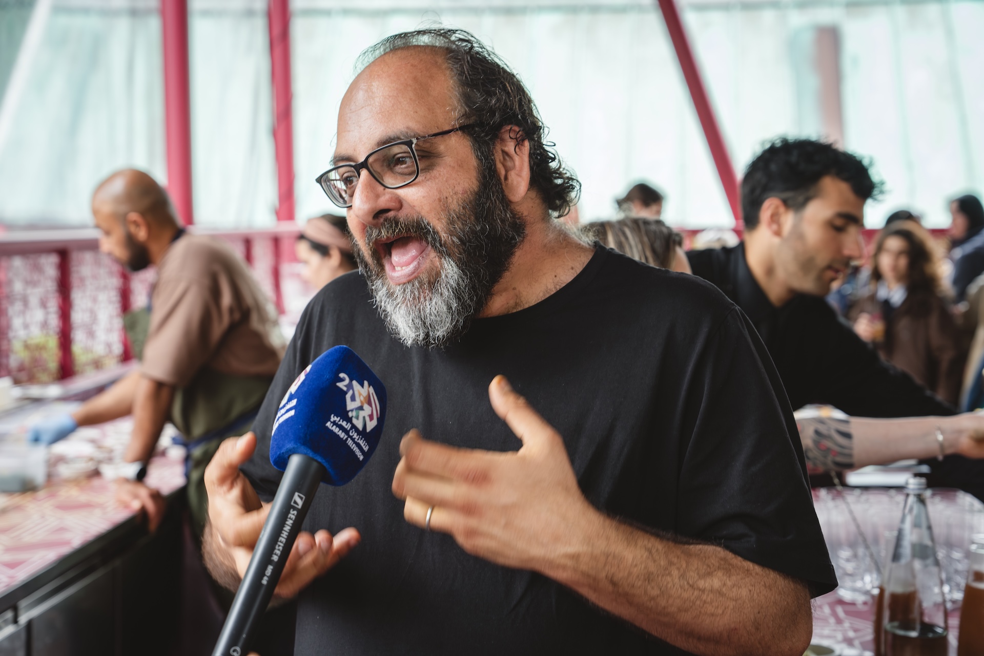 A man with glasses and a beard passionately speaks into a blue microphone labeled “2M” in a busy indoor setting, possibly covering events like the Fashion Trust Arabia Prize, with people socializing and working in the background.