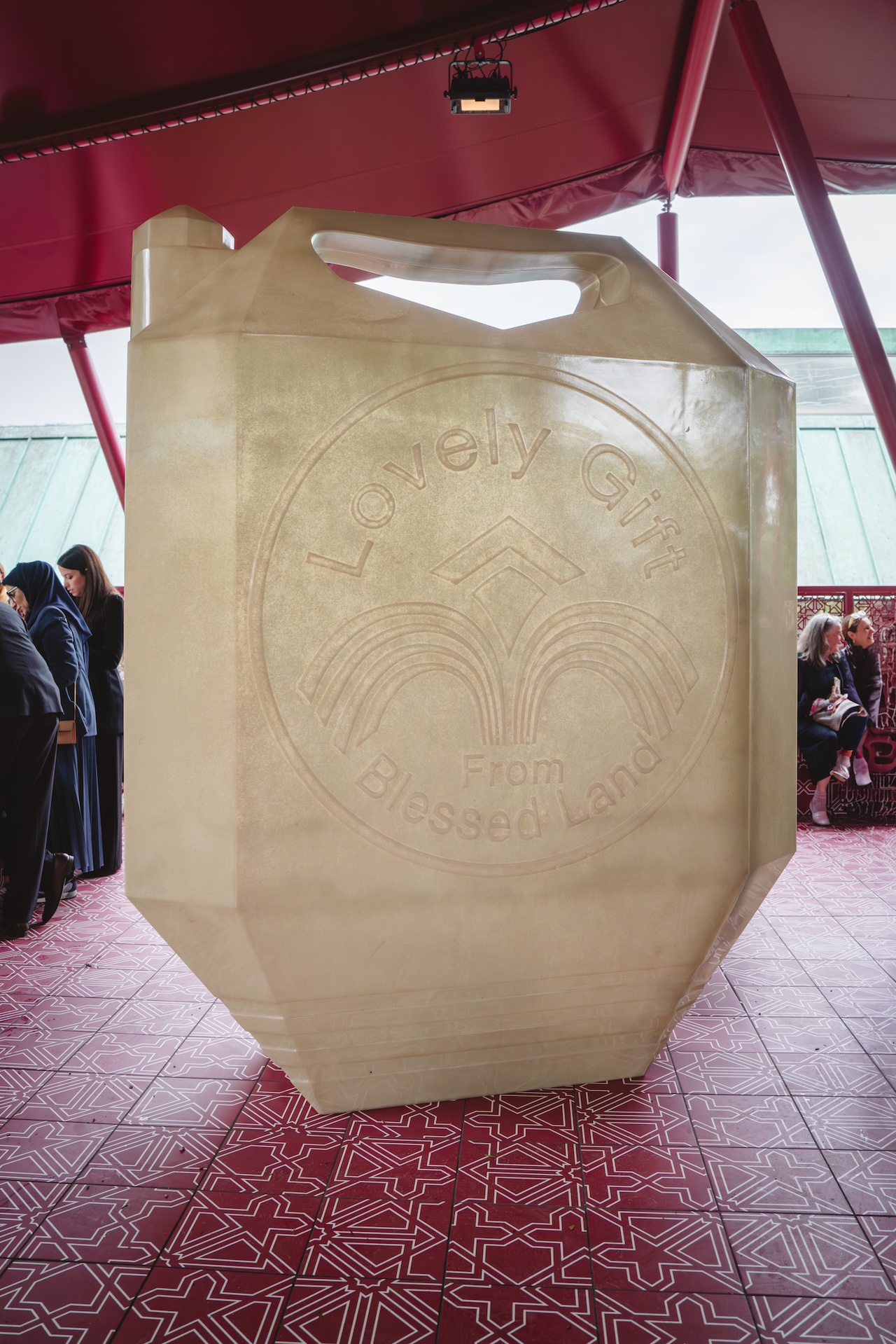 A large translucent sculpture shaped like a jerry can stands indoors, embossed with the words Lovely Gift From Blessed Land. People gather in the background during the Fashion Trust Arabia Prize, and the floor features a red patterned tile.
