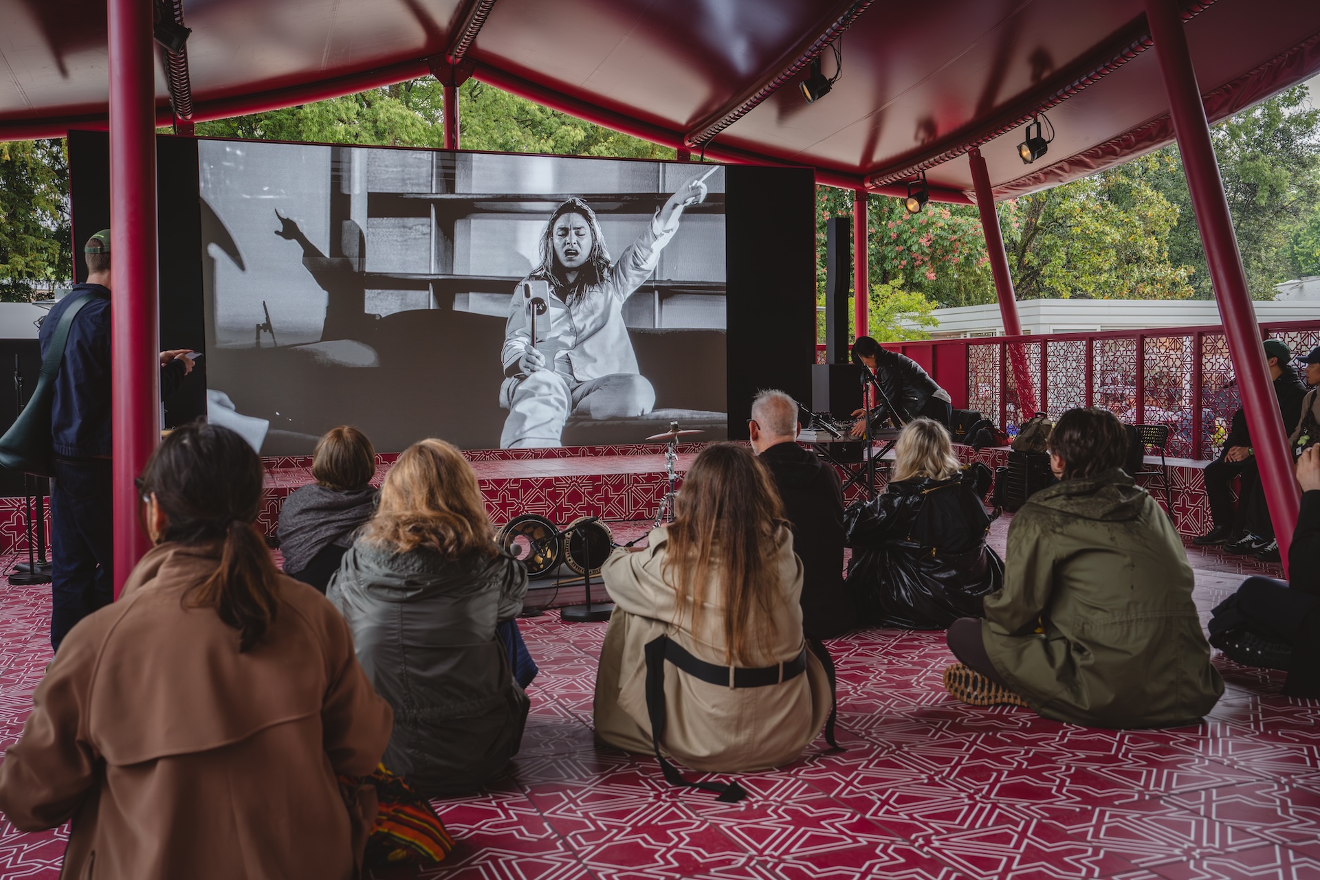 A group of people sit on a patterned floor inside a red pavilion, watching a large black and white screen showing a person speaking and gesturing with one arm raised, possibly during the Fashion Trust Arabia Prize event. Trees are visible outside the pavilion.