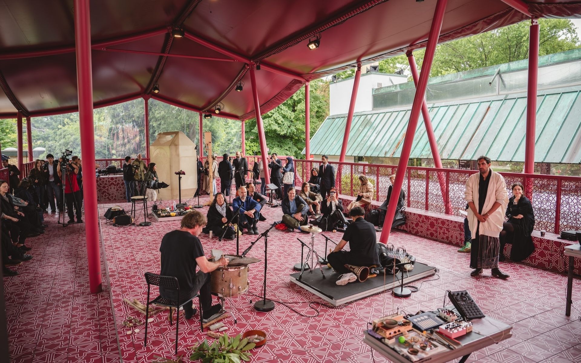 Musicians perform under a red canopy with an audience seated and standing around them. The floor and columns, inspired by Fashion Trust Arabia Prize flair, have matching red patterns, and trees are visible outside. Instruments are scattered on the ground.