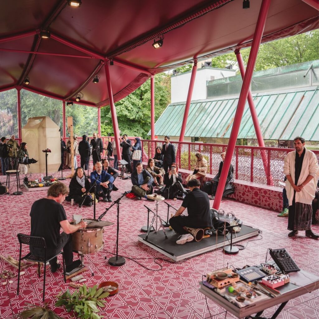 Musicians perform on a patterned red stage under a canopy, with an audience seated and standing in the background. Instruments and plants are visible; trees and buildings are seen outside the open sides.