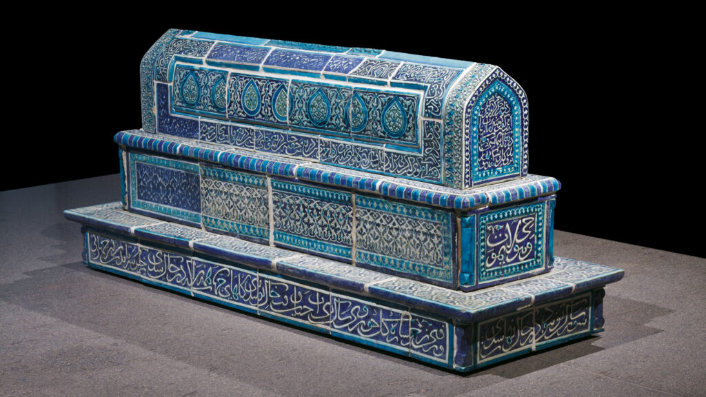 A rectangular, blue-tiled tomb with ornate Islamic calligraphy and geometric patterns, sitting on a stone platform against a dark background.