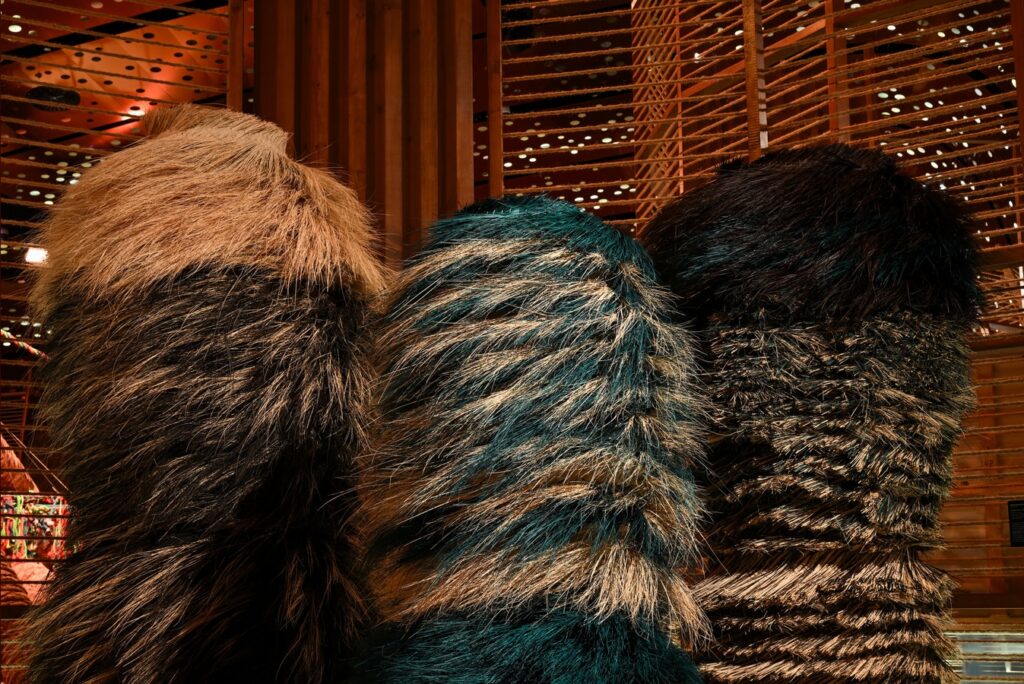 Three tall sculptures covered in layers of textured, fur-like material in shades of brown, teal, and black stand before a wooden latticed wall.