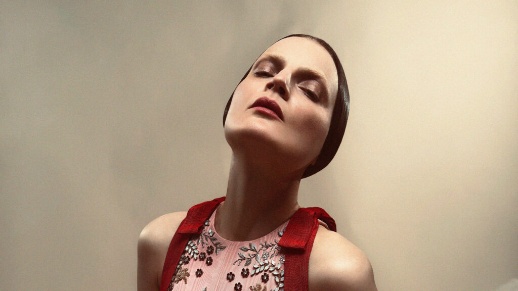 A person with slicked-back dark hair and closed eyes tilts their head back, wearing a pink top with floral embroidery and bold red straps, against a neutral background.
