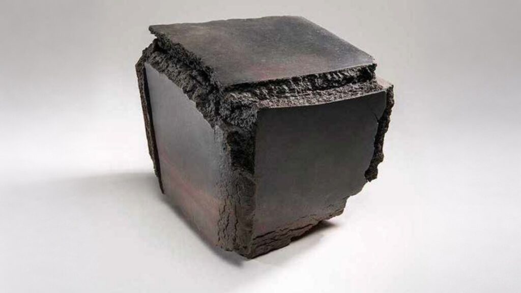 A dark, cube-shaped sculpture with smooth, flat sides and textured, rough edges.