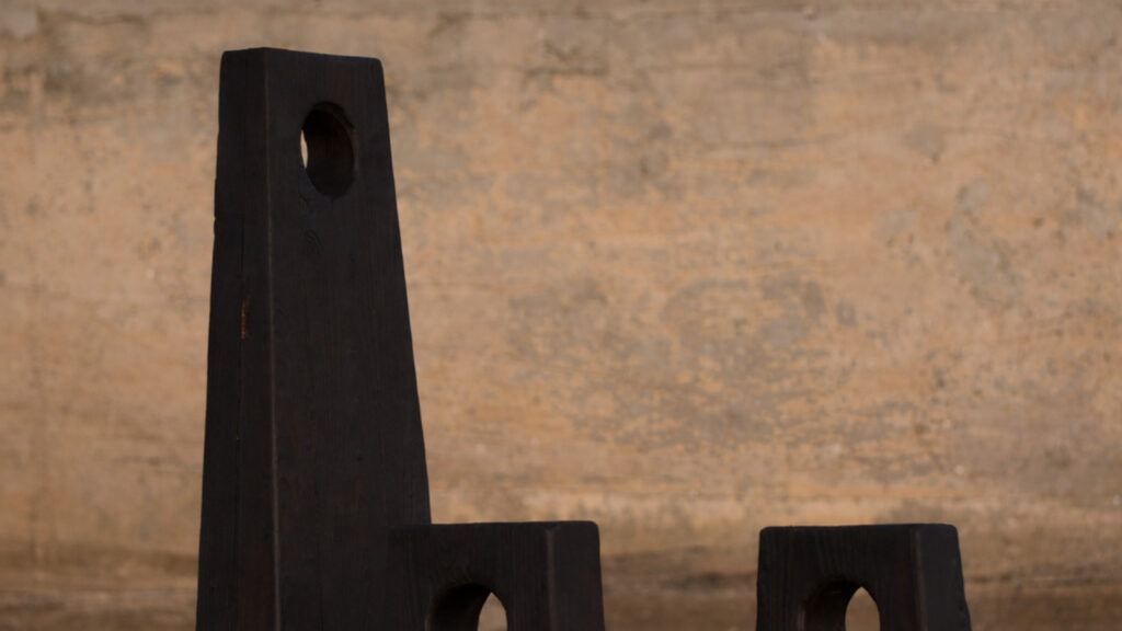 Three dark wooden blocks with circular holes stand in front of a textured beige wall. The blocks are different heights and have a minimalist, geometric design.