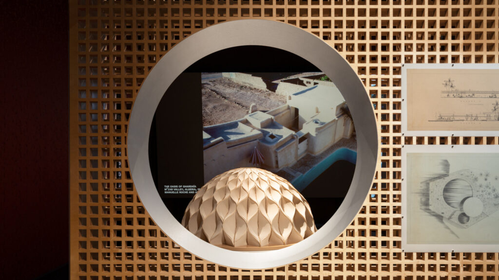 A geometric dome model is displayed in a circular frame, in front of architectural drawings and a photo of tan buildings with angular roofs and a pool.