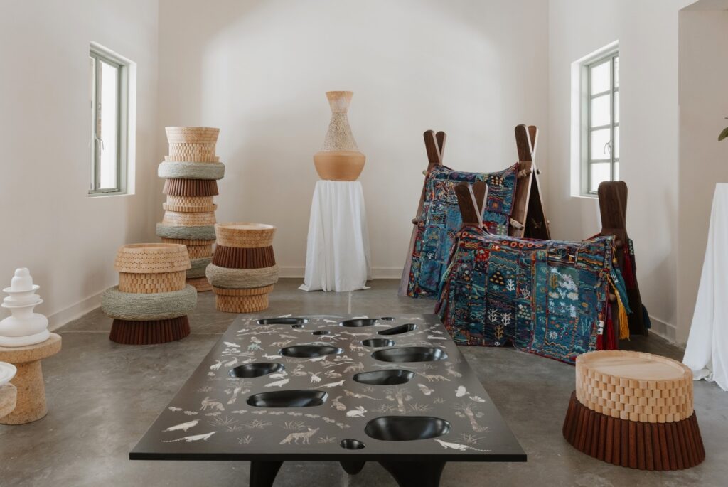 A room featuring a black table with organic-shaped indentations and white animal illustrations, surrounded by woven stools, a tall vase on a pedestal, and textile-covered A-frame structures.