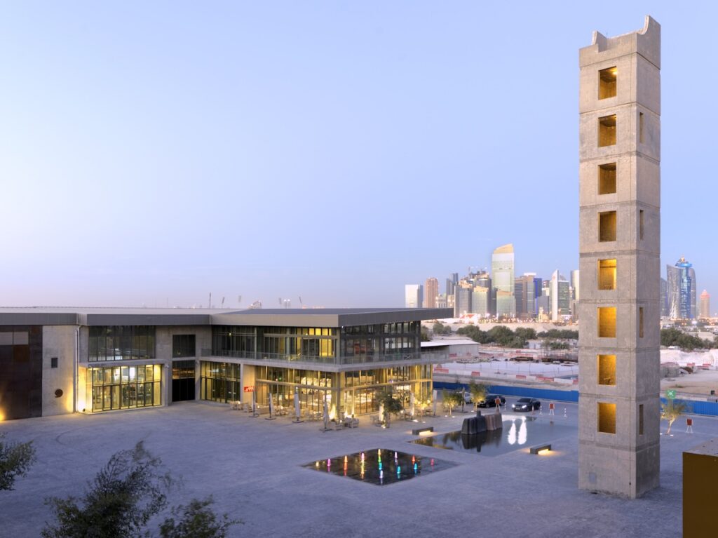 A modern building complex in the style of Rirkrit Tiravanija, featuring a tall, narrow concrete tower, a courtyard with colorful fountain lights, and a distant city skyline at dusk.