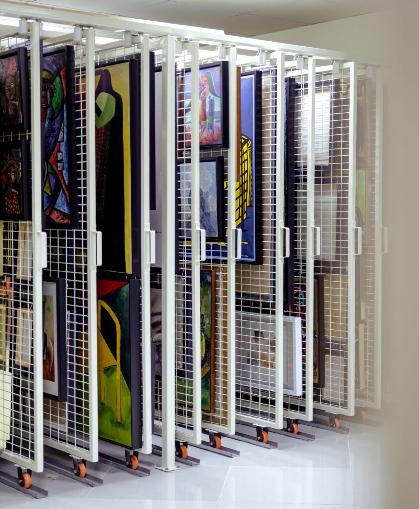 Rows of paintings and artwork are stored upright on metal racks with mesh panels and wheels in a well-lit storage room