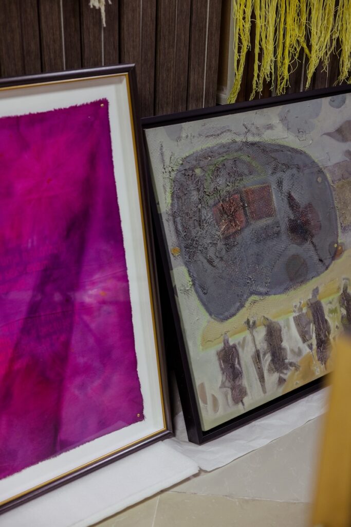 Two framed abstract paintings lean against a wall; one has a vivid pink design, and the other features muted grays and yellows with textured elements