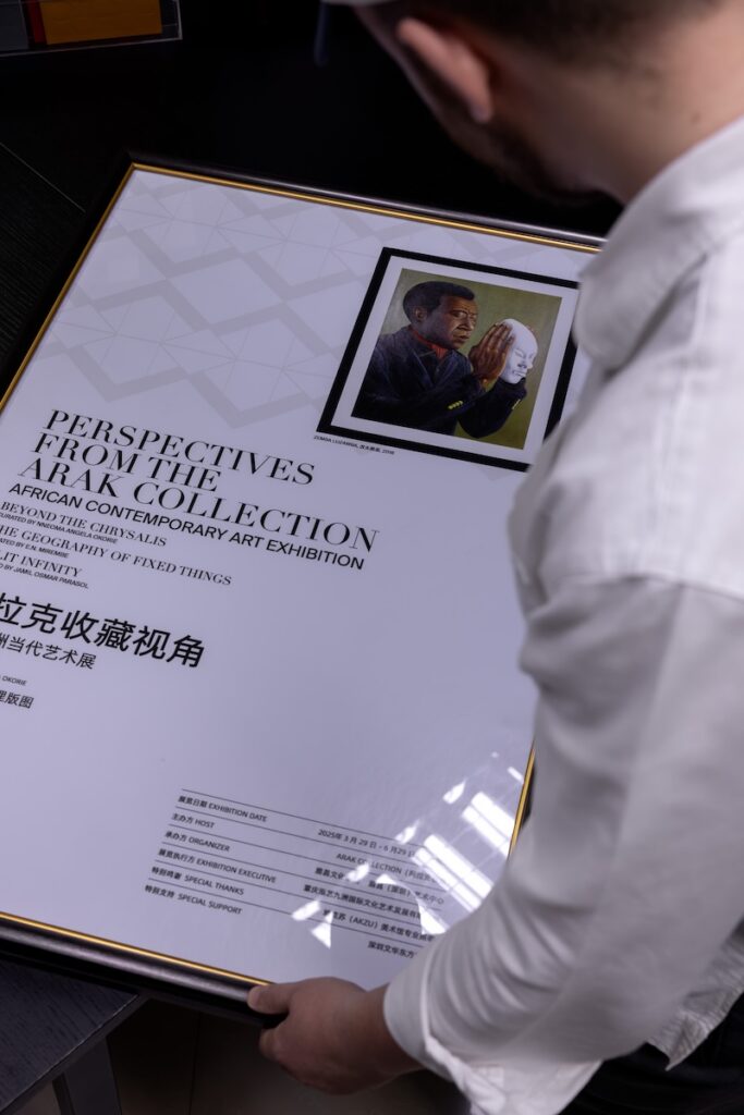 A person holds a framed exhibition poster titled Perspectives from the African Collection, featuring an image of a man holding a white mask. The poster includes English and Chinese text about the art exhibition, and highlights renowned cultural figures such as Chef Noof Al Mari.