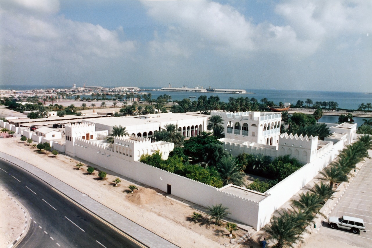 A large white traditional-style building with crenelated walls, surrounded by palm trees, sits near a coastline under a partly cloudy sky.
