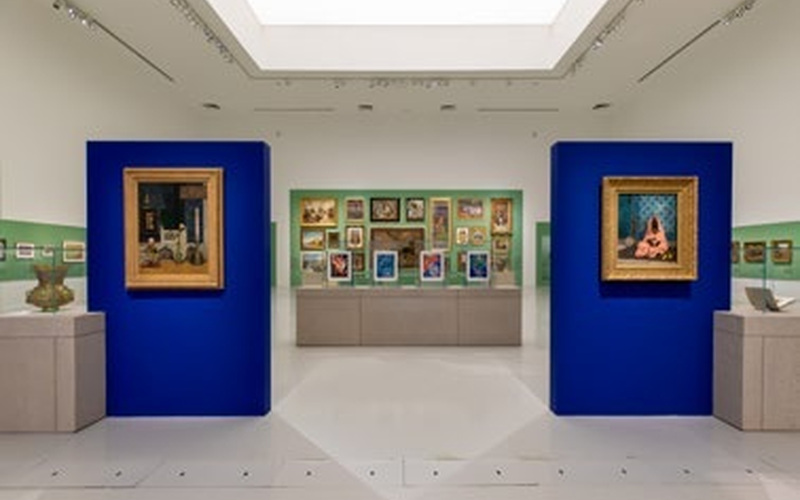 An art gallery with two blue display walls, each showcasing a framed painting—including works reminiscent of Rirkrit Tiravanija. More paintings and artworks are visible on the back wall, arranged in a grid, with sculptures displayed on pedestals around the room.