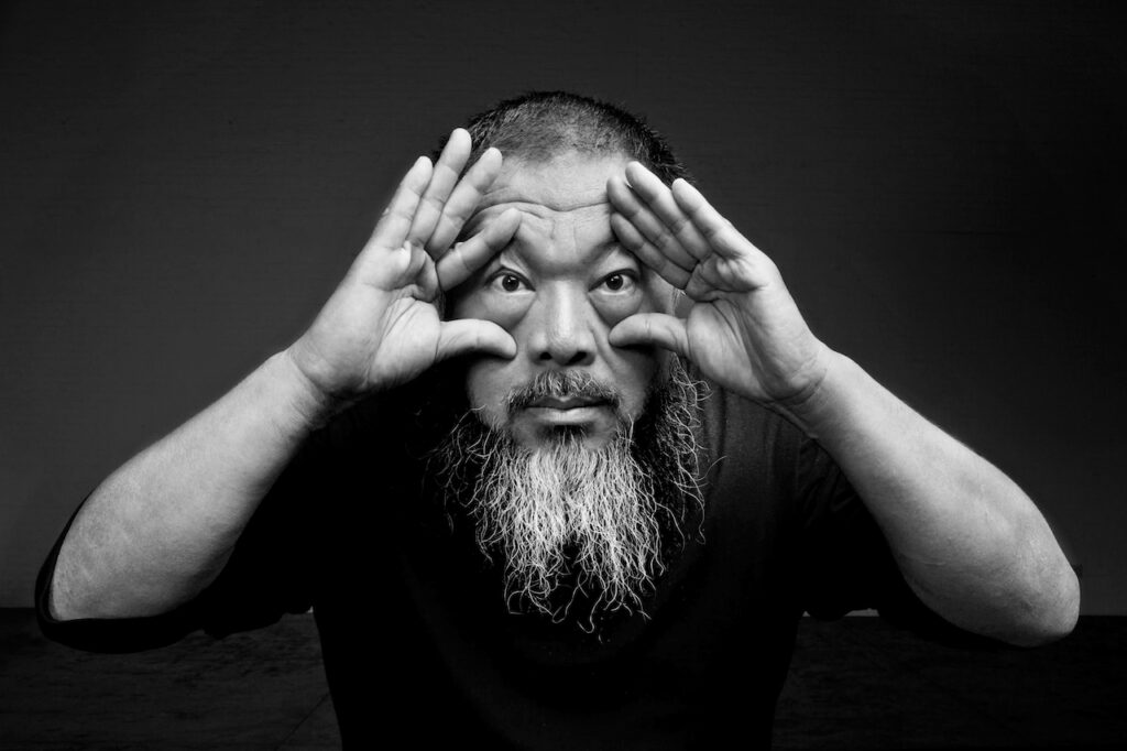 Rirkrit Tiravanija, a middle-aged man with a graying beard, holds his hands to his eyes to frame them as he stares directly at the camera.