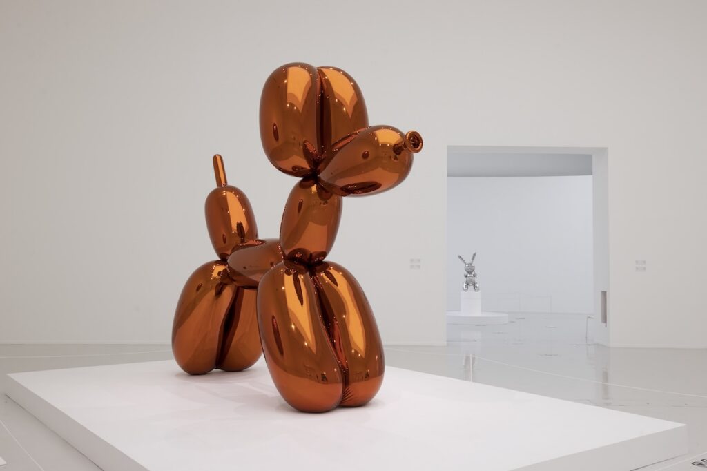 A large, orange, reflective Rirkrit Tiravanija balloon dog sculpture stands on a white pedestal in a gallery, with a smaller silver rabbit sculpture visible in the background.