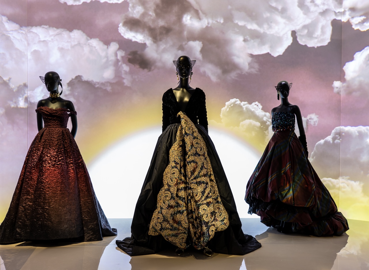 Three mannequins display elegant, dramatic ball gowns in dark colors with textured and patterned fabrics, set against a backdrop featuring clouds and a glowing sun